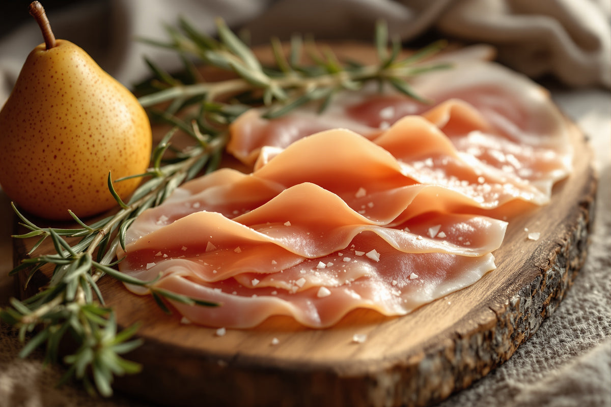 Thin slices of rose-pink prosciutto draped over a wooden board beside a whole pear and sprig of rosemary, soft daylight 