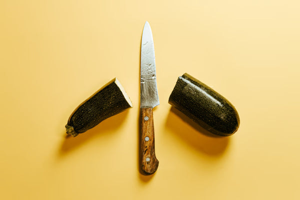 A chef's knife mid-cut through a pile of brunoise-diced shallots on a worn wooden board, soft window light, scattered herb stems.