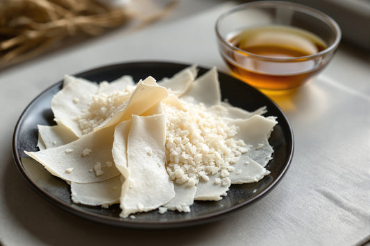 Dried kombu sheets and a mound of pale katsuobushi flakes arranged on a dark ceramic plate beside a glass bowl of clear 