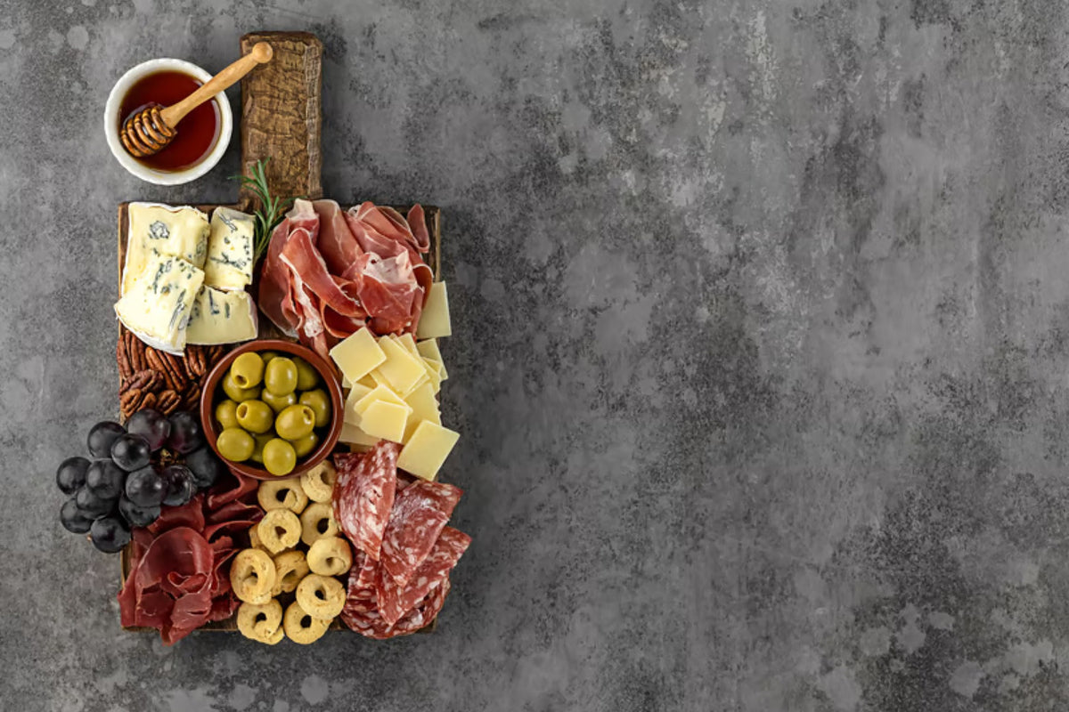 A rustic wooden board with three wedges of cheese, three types of cured meat, three small bowls of olives and grapes, and three crackers arranged in natural daylight.