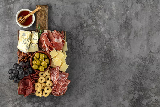 A rustic wooden board with three wedges of cheese, three types of cured meat, three small bowls of olives and grapes, and three crackers arranged in natural daylight.