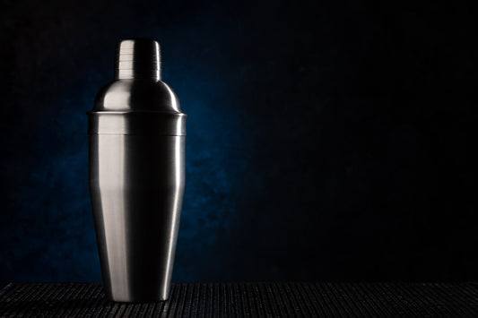 stainless cocktail shaker and boston tin set on dark bar counter with citrus, soft evening light, no text no logos
